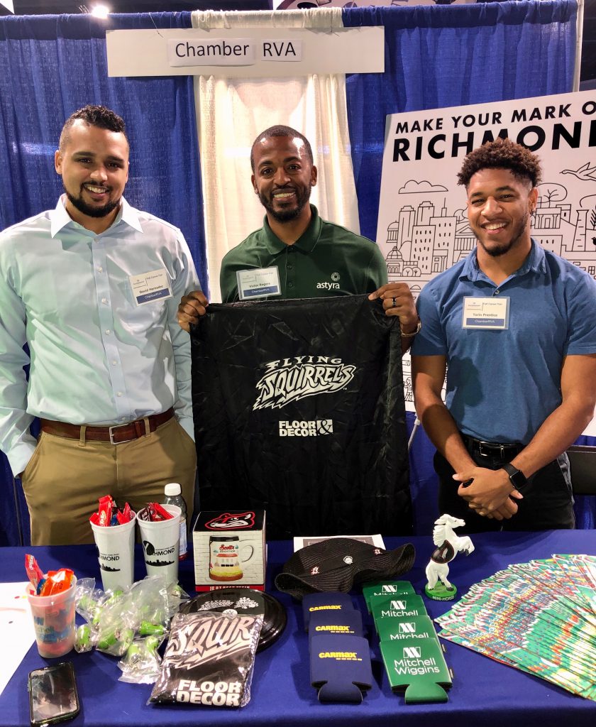 David, Victor and Torin all pose together smiling behind the ChamberRVA table. Victor is holding up a black bag with the Flying Squirrels logo in white.