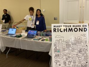 Tianna and Shaila pose together smiling behind the ChamberRVA table. Tianna is extending her arm to bring attention to the different swag on the table.